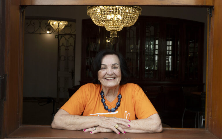 Mulher idosa de cabelos escuros e curtos sorri enquanto apoia os braços no batente de uma janela de madeira. Ela veste camiseta laranja, colar de contas azuis e anéis nas mãos. Ao fundo, no interior iluminado do ambiente, destaca-se um lustre de cristal suspenso no teto.