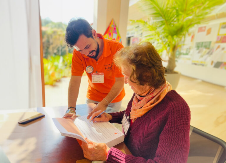 Rapaz com camisa polo laranja do Fiuza inclinado sobre uma mesa, orientando uma mulher idosa de blusa de tricô vinho que escreve em um caderno impresso.