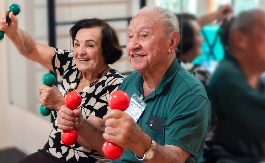 Mulher e homem idosos sorriem alegremente enquanto praticam exercícios físicos segurando pequenos halteres coloridos em uma sala de treino.