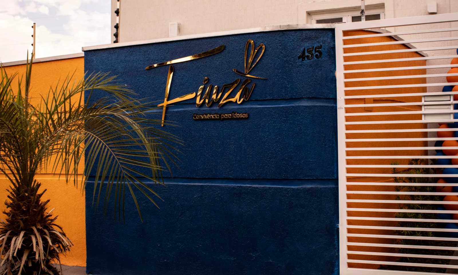 Fachada da nova unidade do Fiuza em Cuiabá, destacando um muro azul escuro com o letreiro dourado "Fiuza - Convivência para Idosos", o número 455 e um portão branco de grade. À esquerda, há um muro amarelo e uma pequena palmeira, e ao fundo, a estrutura do imóvel.