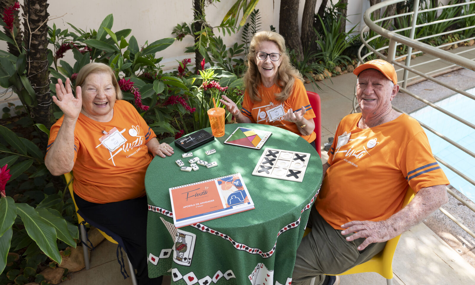 Três idosos sorridentes, vestidos com camisetas laranja do projeto Fiuzão, estão sentados ao redor de uma mesa com jogos recreativos em uma área externa.