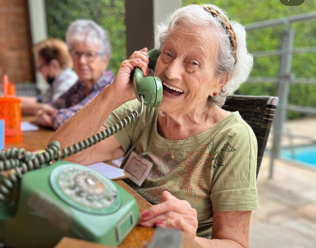Mulher idosa sorridente falando em um telefone antigo de cor verde, sentada à mesa em um espaço de convivência ao ar livre com outras pessoas ao fundo.