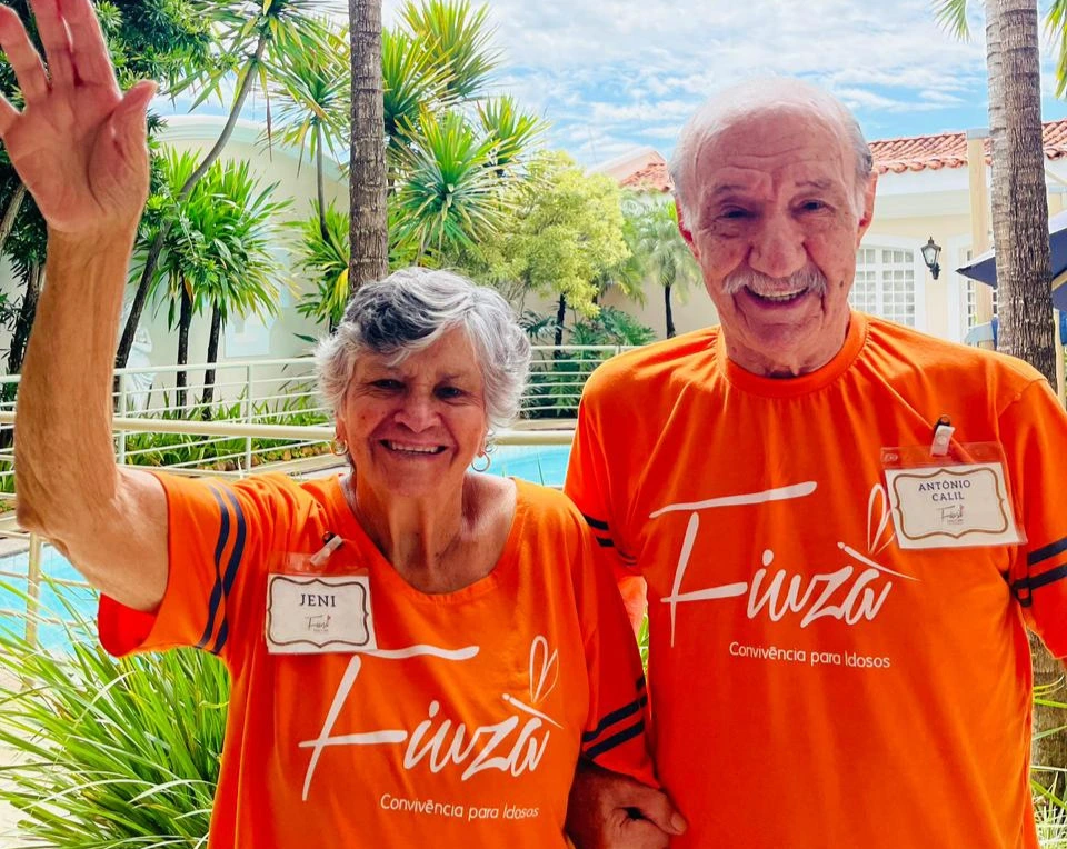 Duas pessoas idosas sorridentes vestem camisetas laranjas do Fiuza em uma área externa arborizada.